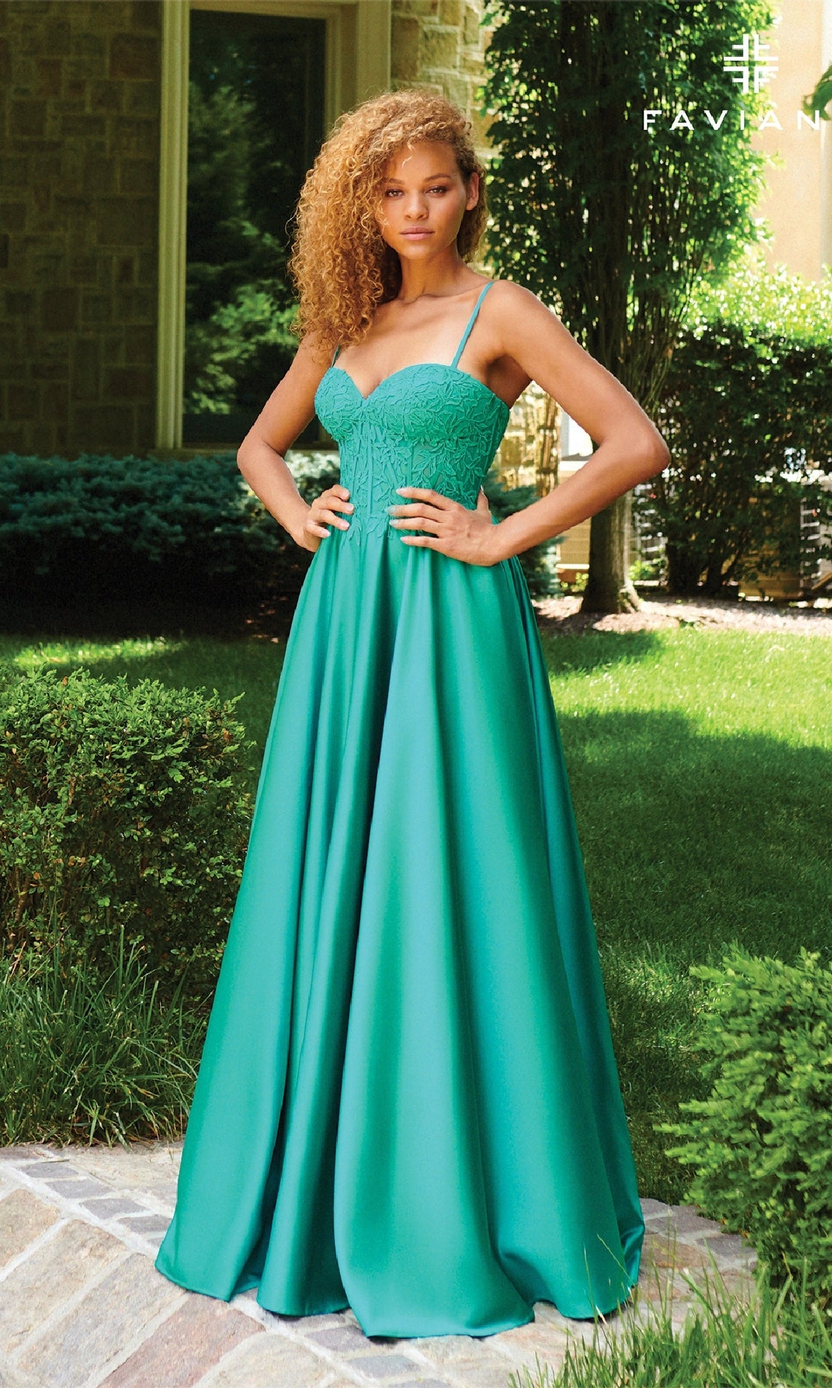 Woman outdoors in the Faviana 11259 Lace-Up Long A-Line Prom Dress, featuring a sweetheart embroidered-lace corset bodice and flared teal skirt, standing with hands on hips in front of greenery and a building.